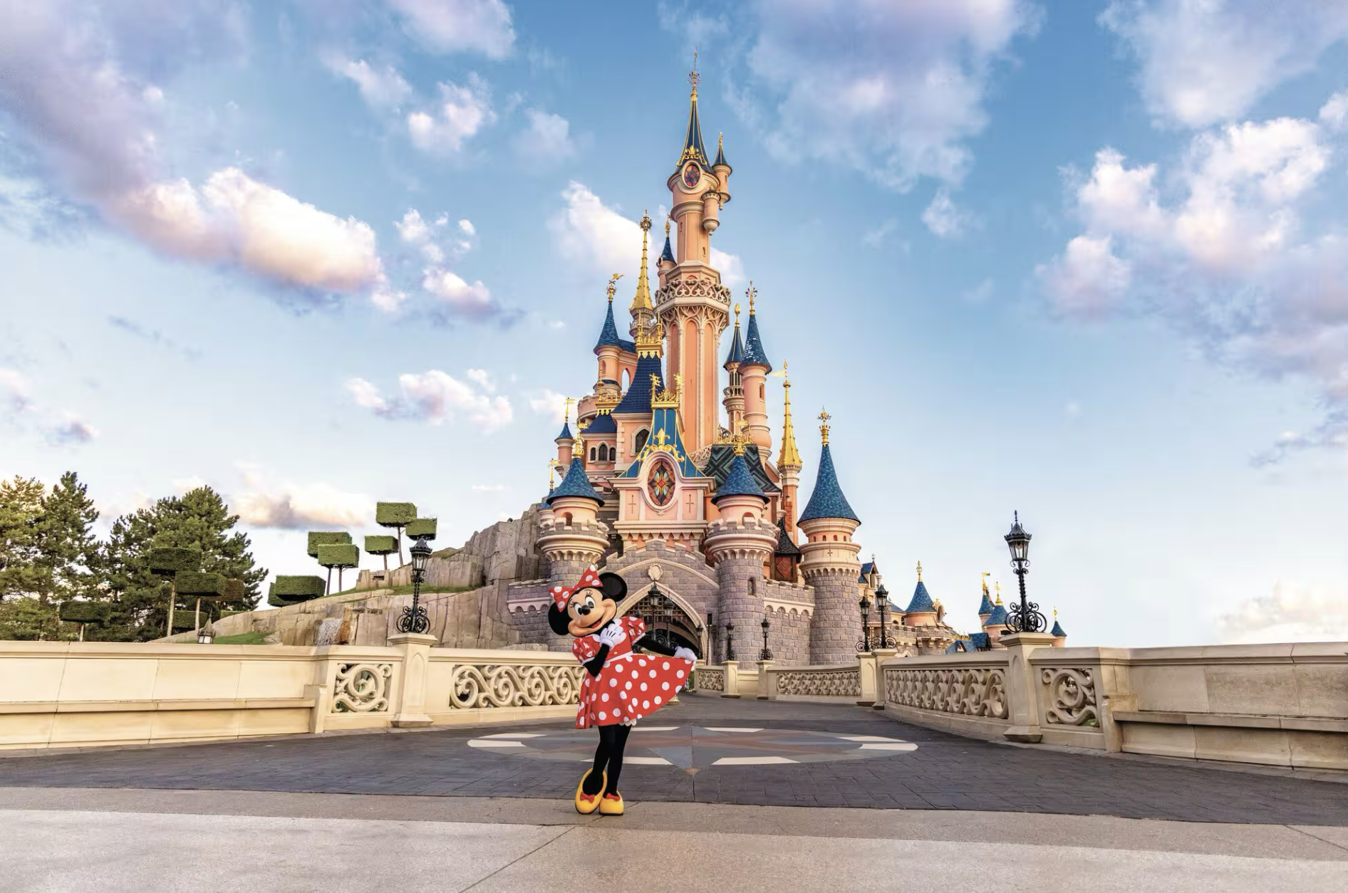 Castillo de la Bella Durmiente en Disneyland París, con Minnie Mouse posando enfrente y un paisaje lleno. de cielo azul, nubes y árboles